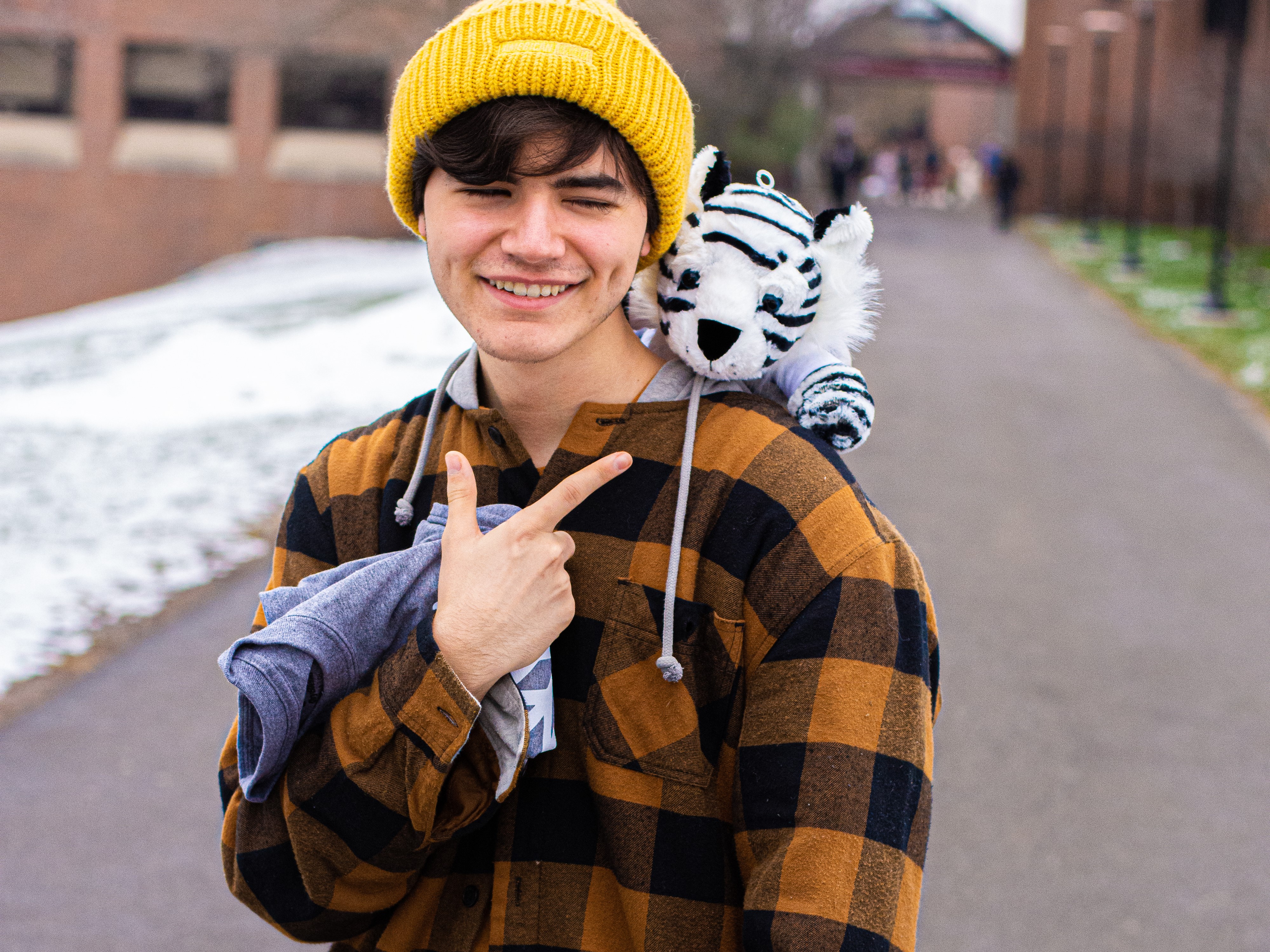 A picture of Nate Papadogiannis in a yellow beaning and orange plaid with a stuffed white tiger on his shoulder. He is pointing at the tiger and smiling.